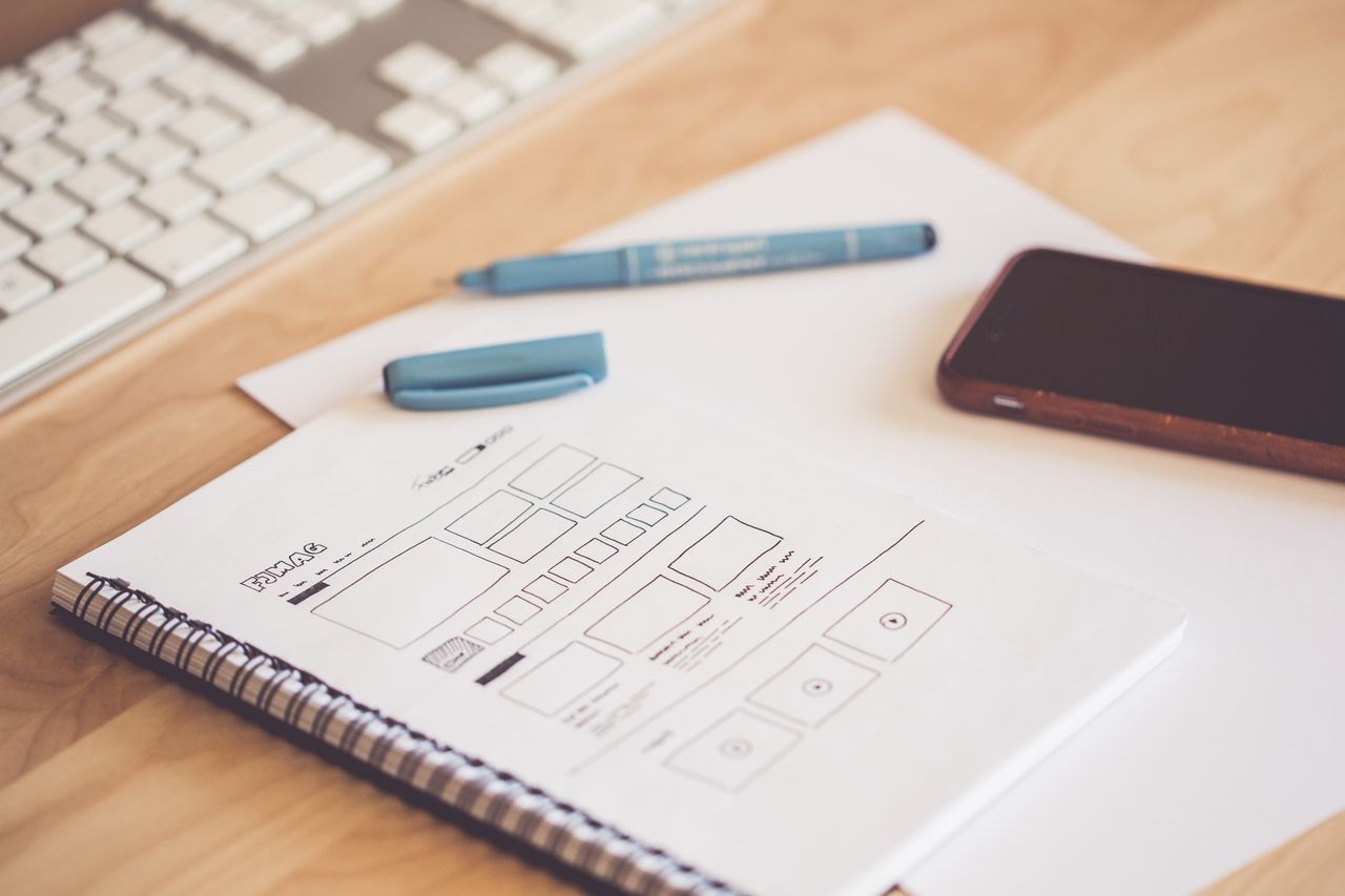 Un espace de travail soigné avec un carnet de croquis contenant des wireframes, un smartphone et un clavier posés sur un bureau en bois.